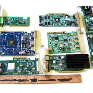 May include: A collection of seven assorted computer expansion cards, including green and blue printed circuit boards with various electronic components, heat sinks, and metal brackets, arranged above a 12-inch wooden ruler for scale.