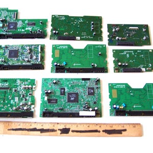 Op de afbeelding: Een verzameling groene printplaten met diverse elektronische componenten, waaronder chips, connectoren en weerstanden. Een houten liniaal met inch-markeringen staat onderaan. De platen variëren in grootte en vorm.