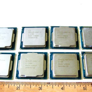 May include: Eight square computer processors arranged in two rows of four, each featuring a silver metal top plate with printed text and a dark green circuit board base, positioned above a wooden ruler measuring approximately 6.5 inches in length.