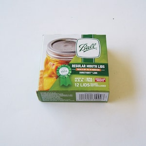 May include: A box of 12 Ball regular mouth canning lids, made in the USA. The box features a green and white design with the Ball logo and the text "Regular Mouth Lids" and "SureTight" lids. The box also states that the lids are BPA free and that bands are not included.