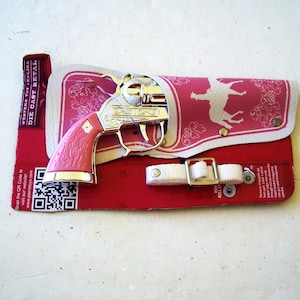 Op de afbeelding: Een speelgoed replica van een Western pistool van gegoten metaal met een roze handvat en zilveren accenten, rustend op een rode en roze holster met een witte riem. De holster heeft een wit silhouet van een paard.