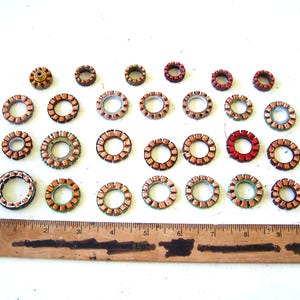 May include: A collection of twenty-five circular copper-wound motor stators of varying sizes arranged in four rows. A wooden ruler is positioned at the bottom, showing the largest components measure approximately 1.5 inches in diameter.