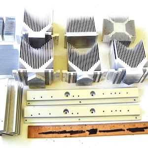 May include: A collection of silver-colored aluminum heat sinks with various fin configurations and two long, flat mounting bars, arranged alongside a wooden ruler that measures 12 inches in length to provide a clear scale for the components.