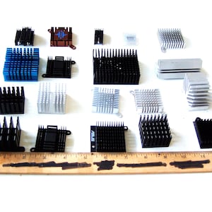May include: A selection of computer heat sinks in various shapes, sizes, and colours, including black, blue, silver, and brown. A wooden ruler is placed at the bottom of the image, with measurements in inches and centimetres.