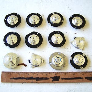 May include: A collection of twelve circular metal and black plastic motor components arranged in three rows, positioned above a wooden ruler that measures approximately 11 inches in length, showing various mechanical parts with central hubs.