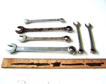 May include: A collection of six metal combination wrenches of varying sizes, including some with visible rust and others with a polished finish, arranged above a wooden ruler that measures up to 12 inches in length for scale reference.