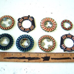 May include: Eight circular electric motor stators with copper or dark blue wire windings arranged in two rows above a wooden ruler that measures 10 inches in length. The components feature metal cores and various mounting points for assembly.