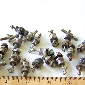 May include: A collection of 20 silver-toned diodes with black and red markings. The diodes are arranged in a random pattern on a white surface. A wooden ruler with inch markings is visible at the bottom of the image.