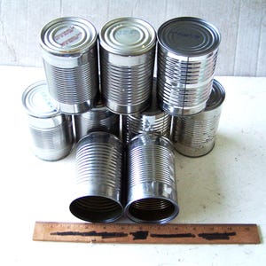 May include: A collection of ten empty, cylindrical metal food cans with ribbed sides and flat lids, arranged in a group. A wooden ruler is placed horizontally in front of the cans, showing a length of approximately 12 inches.