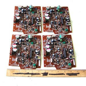 May include: Four brown Toshiba MKT3-MAINS circuit boards with various electronic components including capacitors, resistors, and integrated circuits. A wooden ruler is placed along the bottom edge, showing a width of approximately 12 inches.