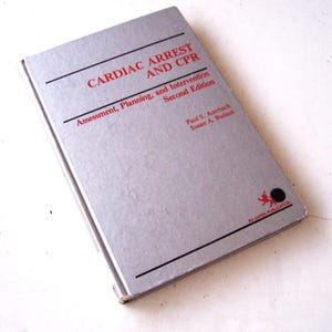 May include: A silver book titled "CARDIAC ARREST AND CPR" with red text. The book's subtitle reads "Assessment, Planning, and Intervention, Second Edition." The cover also includes the authors' names and the Aspen Publication logo.