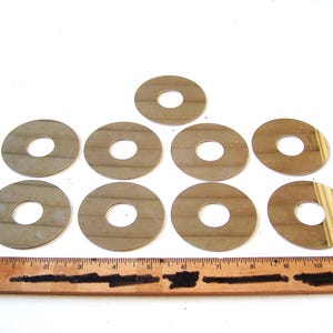 Könnte beinhalten: Zehn kreisförmige Metallscheiben mit einem zentralen Loch, angeordnet auf einer weißen Oberfläche. Ein Holzlineal mit schwarzen Markierungen ist darunter platziert. Die Scheiben haben einen metallischen Glanz.