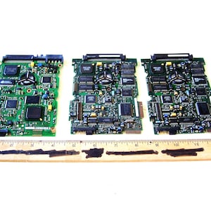 Puede incluir: Tres placas de circuito impreso verdes con varios componentes electrónicos, incluyendo chips y conectores. Una regla de madera con marcas en centímetros se coloca debajo. Las placas están dispuestas una al lado de la otra.