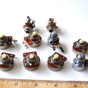 Könnte beinhalten: Eine Sammlung elektronischer Potentiometer mit verschiedenen Größen und Designs. Diese Komponenten haben einen silberfarbenen Metallkörper, einen cremefarbenen Knopf und eine rotbraune Basis. Ein Holzlineal befindet sich im Vordergrund.