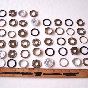 May include: An assortment of metal washers and black rubber O-rings are arranged on a white surface. The washers vary in size and design, with some having a silver finish and others white. A wooden ruler is at the bottom, providing scale.