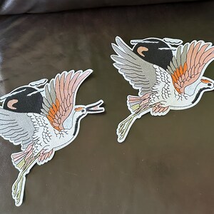 May include: Two embroidered iron-on patches of cranes in flight. The cranes are white with black, gray, orange, and pink accents. The patches are perfect for adding a touch of nature to clothing, bags, or other fabric items.