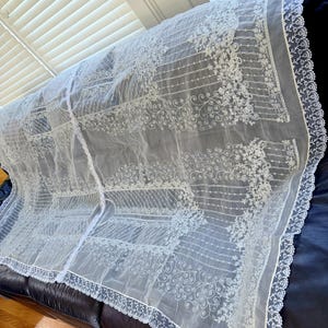 May include: A sheer white lace curtain panel featuring intricate floral embroidery and scrolling vine patterns. The fabric has a delicate mesh texture with a scalloped lace border along the edges, showcasing a repeating decorative design.