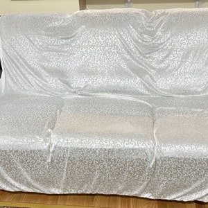 May include: A white couch cover with a delicate swirl pattern. The cover is made of a soft, silky fabric and is draped over a black leather sofa.