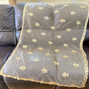 May include: A sheer, pale scarf with gold-coloured embellishments. The edges are trimmed with a gold border and small, white beads. The scarf is draped over a dark leather surface.