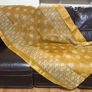 May include: A yellow and white floral patterned fabric draped over a brown leather couch. The fabric has a gold border.