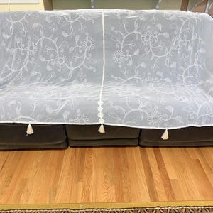 May include: White sheer sofa cover with embroidered floral design. The cover has a delicate pattern of flowers and vines, with tassels at the corners and along the bottom edge. It is draped over a dark brown sofa.