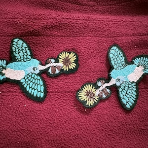 May include: Two embroidered blue hummingbird patches with pink and white accents. Each hummingbird is depicted with a long beak and is shown drinking nectar from a yellow flower with brown and white accents.