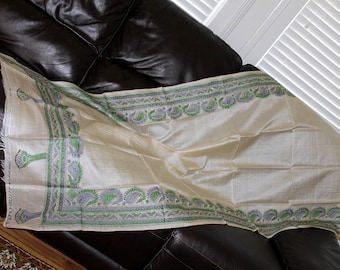 May include: A white silk scarf with a green and blue paisley border. The scarf is folded and lying on a brown leather couch.
