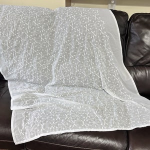 May include: A white, sheer fabric with an embroidered floral pattern is draped over a dark brown leather sofa. The delicate embroidery creates a textured surface, adding visual interest to the textile.