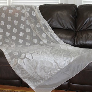 May include: A white sheer curtain with a geometric pattern of squares and diamonds. The curtain is draped over a brown leather sofa.