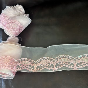 May include: Pink lace trim with a scalloped edge and a delicate floral design. The lace is on a sheer white fabric.