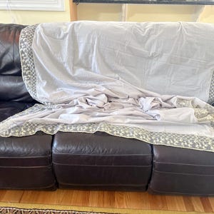 May include: A white crocheted throw blanket with a delicate floral pattern and tassel fringe draped over a brown leather sofa.