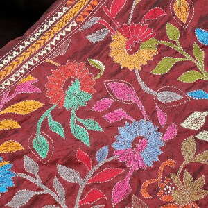 May include: Burgundy fabric with colorful floral embroidery. The design features flowers in shades of red, orange, blue, and pink, with green and gray leaves. The edges have a decorative border.