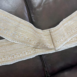 May include: Cream-colored embroidered fabric trim with gold thread detailing. The trim features intricate floral and geometric patterns. This decorative trim is suitable for various sewing and craft projects.