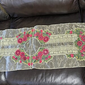 May include: A sheer white fabric table runner with embroidered floral designs in shades of pink, green, and gold. The runner is decorated with a repeating pattern of flowers and leaves.