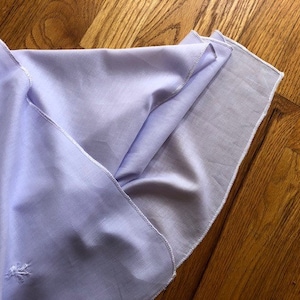 May include: A light purple fabric with a white embroidered design. The fabric is folded and has a white seam around the edges.