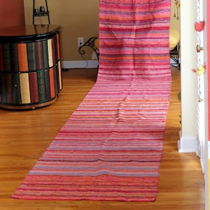 May include: A long, narrow, pink and orange striped rug with a woven texture. The rug is laid out on a wooden floor.