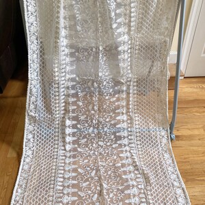 May include: A white sheer fabric with intricate embroidery in a floral pattern. The fabric has a scalloped edge and a white pom-pom trim.