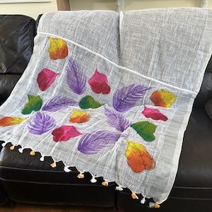 May include: White linen scarf with hand-painted colorful leaves in shades of purple, green, yellow, orange, and pink. The scarf has white and yellow tassels.