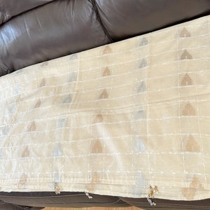 May include: A cream-colored sheer curtain panel with a subtle geometric pattern in shades of brown and silver. The curtain has a beaded fringe along the bottom edge.