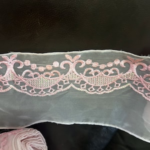 May include: Pink embroidered lace trim with a delicate floral pattern on a sheer white fabric.