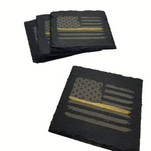 May include: A set of four square black slate coasters featuring a distressed American flag design. Each coaster displays a dark gray flag with a single horizontal gold stripe running through the center, etched onto the textured stone surface.