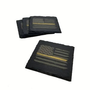 May include: A set of four square black slate coasters with rough, natural edges. Each coaster features a laser-engraved American flag design with a single horizontal gold-colored stripe running through the center of the stripes.