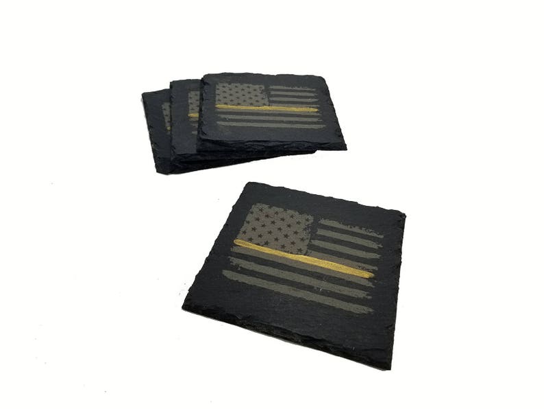 May include: A set of four square black slate coasters with rough, natural edges. Each coaster features a laser-engraved American flag design with a single horizontal gold-colored stripe running through the center of the stripes.