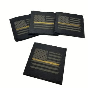 May include: A set of four square black slate coasters featuring a distressed American flag design. Each coaster displays a gray flag with a single horizontal gold stripe running across the center, set against the natural dark textured stone surface.