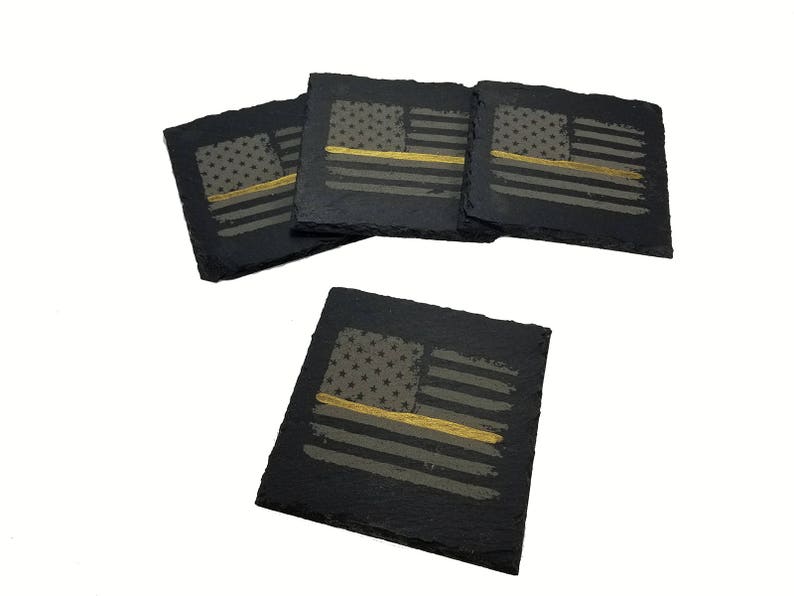 May include: A set of four square black slate coasters featuring a distressed American flag design. Each coaster displays a gray flag with a single horizontal gold stripe running across the center, set against the natural dark textured stone surface.