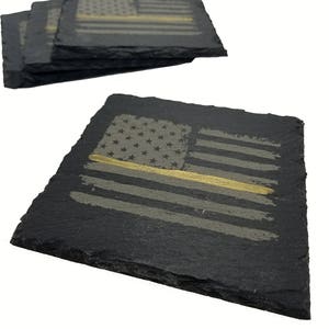 May include: A set of square black slate coasters featuring a laser-engraved American flag design. Each coaster displays a distressed flag pattern with a single horizontal gold line running across the center, highlighting the natural rough edges.
