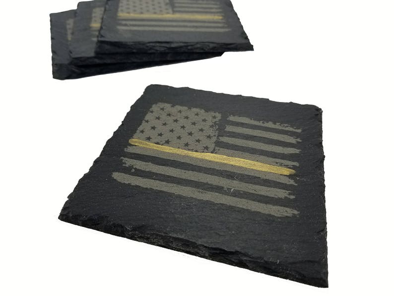 May include: A set of square black slate coasters featuring a laser-engraved American flag design. Each coaster displays a distressed flag pattern with a single horizontal gold line running across the center, highlighting the natural rough edges.