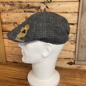 Könnte beinhalten: Eine graue und braune karierte Newsboy-Mütze mit einem gestickten Umriss von Irland und einem Kleeblatt. Die Mütze sitzt auf einem weißen Mannequin-Kopf.