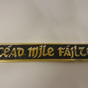 May include: A rectangular metal wall plaque featuring a black textured background with polished gold-coloured raised lettering that reads "CÉAD MÍLE FÁILTE". The plaque has decorative scrollwork at both ends and includes a small hole for mounting.