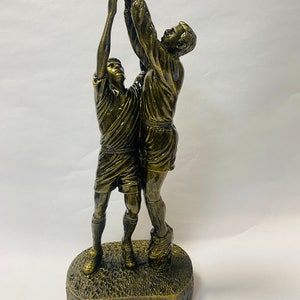 Può includere: Un trofeo color bronzo che raffigura due figure in una posa dinamica, una delle quali alza la mano per prendere una palla mentre l'altra salta per aiutarla. Il trofeo ha una base testurizzata che ricorda l'erba.